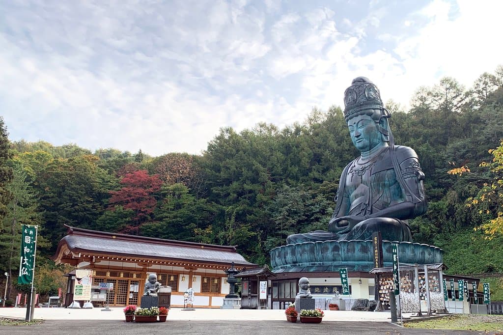 青森市郊外、桑原の地に横たわる稲山は、春には山桜と新緑がみごとな彩りをなし秋には錦を織りなす。まるで天然の屏風のよう。その稲山の眼下には日本の原風景を思わせる田園風景が広がっています。
日本最大の青銅坐像仏、昭和大仏はこの豊かな自然に囲まれた稲山の中腹に鎮座しています。境内地には東日本最大の木造五重塔が屹立し、金堂、開山堂、大師堂、書院が甍を並べ、広大な枯山水庭園「忘帰庭」が訪れる人に落ち着きを与えてくれます。青龍寺伽藍の多くは天然青森ひばを資材に用い、青森の名工、大室勝四郎棟梁が持てる技量を尽くしました。
「伽藍が無言の説法をする」開基織田隆弘師の熱い信念がこの霊場を開き、今もなお全山を満たしています。
日々の喧噪を離れた心静かな時を求めて、国内はもとより世界各国から参拝者が訪れています。