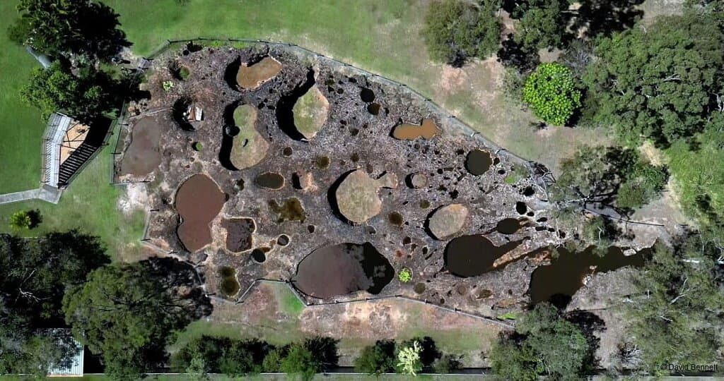 Mystery Craters, seen from above, reveal a captivating mosaic of ochre and green formations set against lush surroundings. This aerial view highlights their ancient allure, with sunlight and shadows adding to the enigma of these geological wonders in Bundaberg