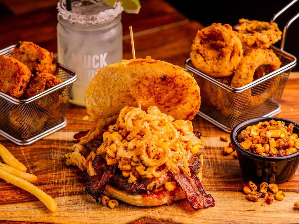The Street Cred burger, featuring a savory Wagyu patty topped with smoky bacon, crispy fried onion strings, and flavorful Mexican street corn, all served on a perfectly toasted bun. Paired with crispy golden onion rings, jalapeño cheddar tots, and a side of roasted street corn, this plate delivers bold flavors and satisfying crunch. Complemented by a refreshing Muck-a-Rita, garnished with a salted rim and lime wedge, this meal is the ultimate indulgence.